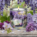 Candle labeled 'Petals on the Path' surrounded by purple flowers on a wooden surface