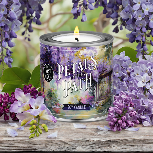Candle labeled 'Petals on the Path' surrounded by purple flowers on a wooden surface