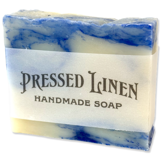 A rectangular bar of handmade soap with a white paper label reading 'PRESSED LINEN HANDMADE SOAP.' The soap has a marbled pattern of blue and cream, with blue bands visible near the top and bottom edges.