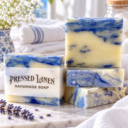 Four handmade Pressed Linen soap bars with a blue and cream marble pattern, stacked on a marble surface with a white cloth, lavender sprigs, and folded towels in the background.