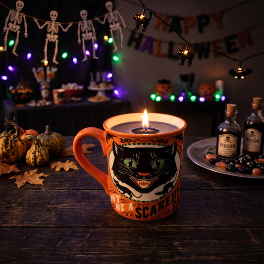 A Halloween-themed mug candle with a lit flame, featuring a black cat illustration and the text 'Don't Be A Scaredy Cat' on an orange ceramic mug, set on a dark wooden table with pumpkins, string lights, and spooky decorations in the background.
