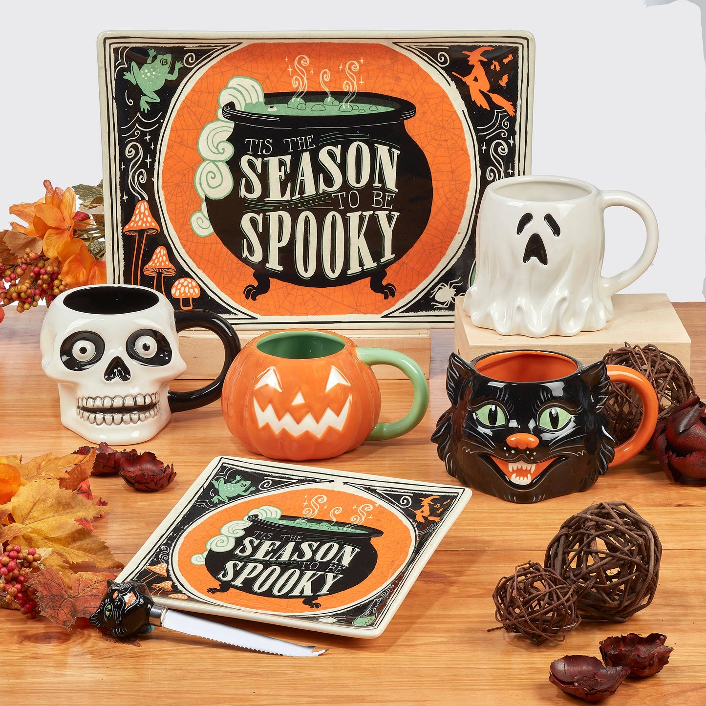 A collection of Halloween-themed ceramic mugs and a matching serving tray arranged on a wooden table with autumn leaves and decorative balls. The set includes a skull mug, a jack-o'-lantern mug, a ghost mug, a black cat mug, and a cauldron mug, alongside a large sign and a smaller square sign that read 'It's the Season to be Spooky' and 'Season Spooky'.
