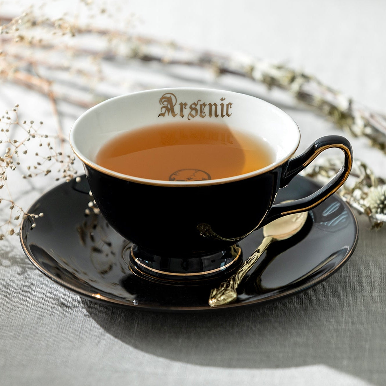 A black ceramic teacup and saucer set with gold trim, filled with tea, on a light gray textured surface with dried flowers and a small spoon. The inside of the cup is white with the word 'Arsenic' printed in gold.
