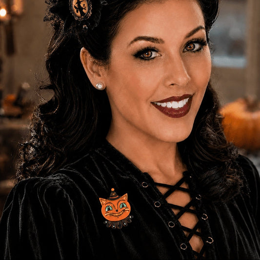 A woman with vintage-style curled dark hair wears a black lace-up velvet top and a small orange pumpkin cat face brooch on the chest. The background is a softly blurred indoor setting with warm candlelight.