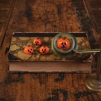 Three small orange jack-o'-lantern-shaped items with black facial features are arranged on a vintage book on a rustic wooden table, with one item viewed through a magnifying glass held by a brass stand.