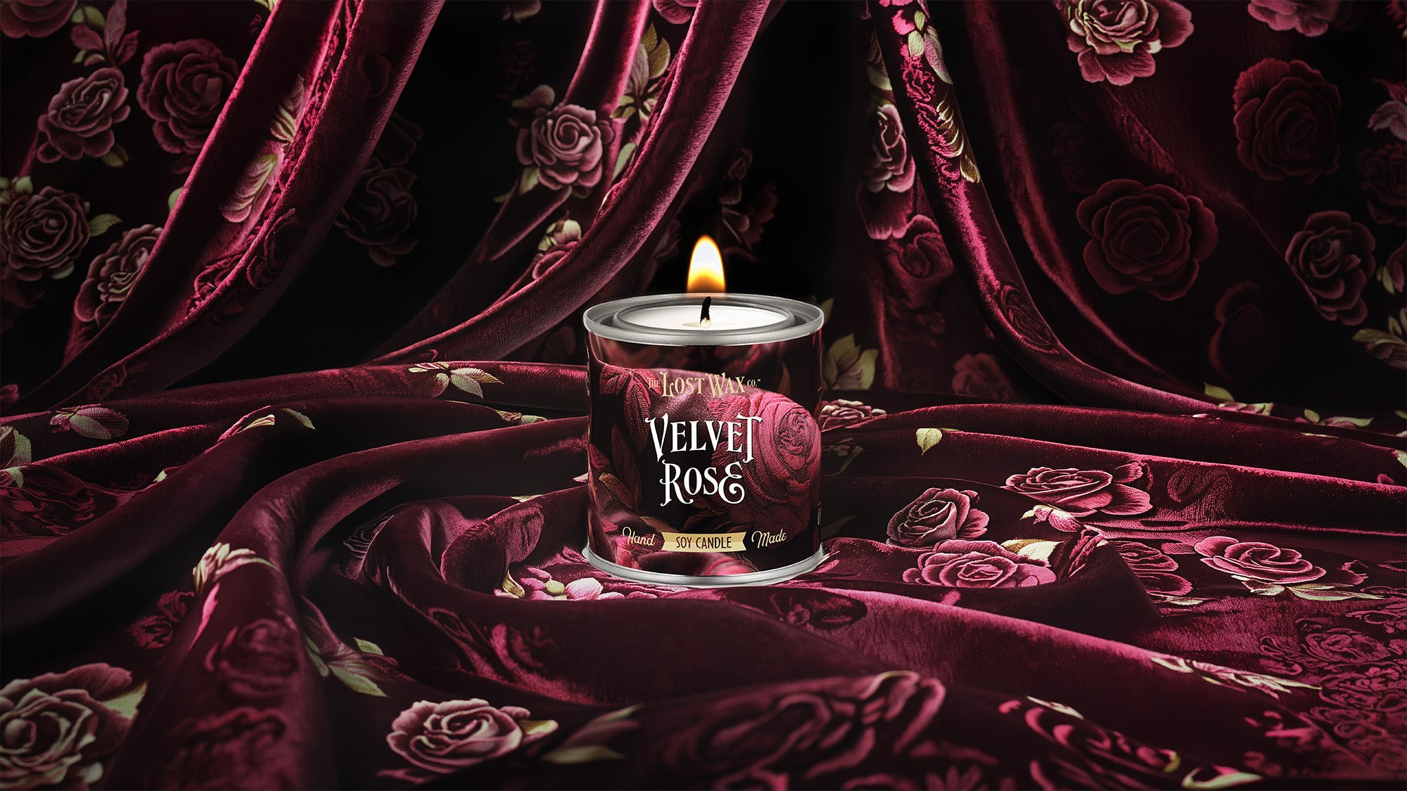 A candle in decorative tin sits surrounded by deep burgundy rose-patterned velvet fabric.