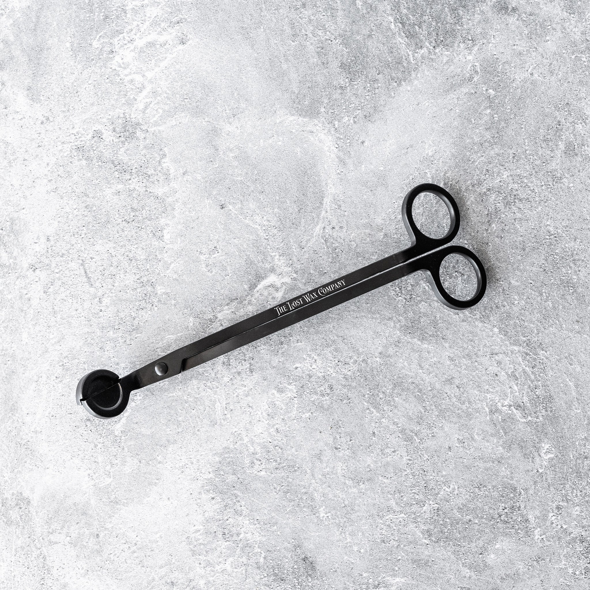 Black metal wick-trimming tool with The Lost Wax Company branding on a textured gray surface