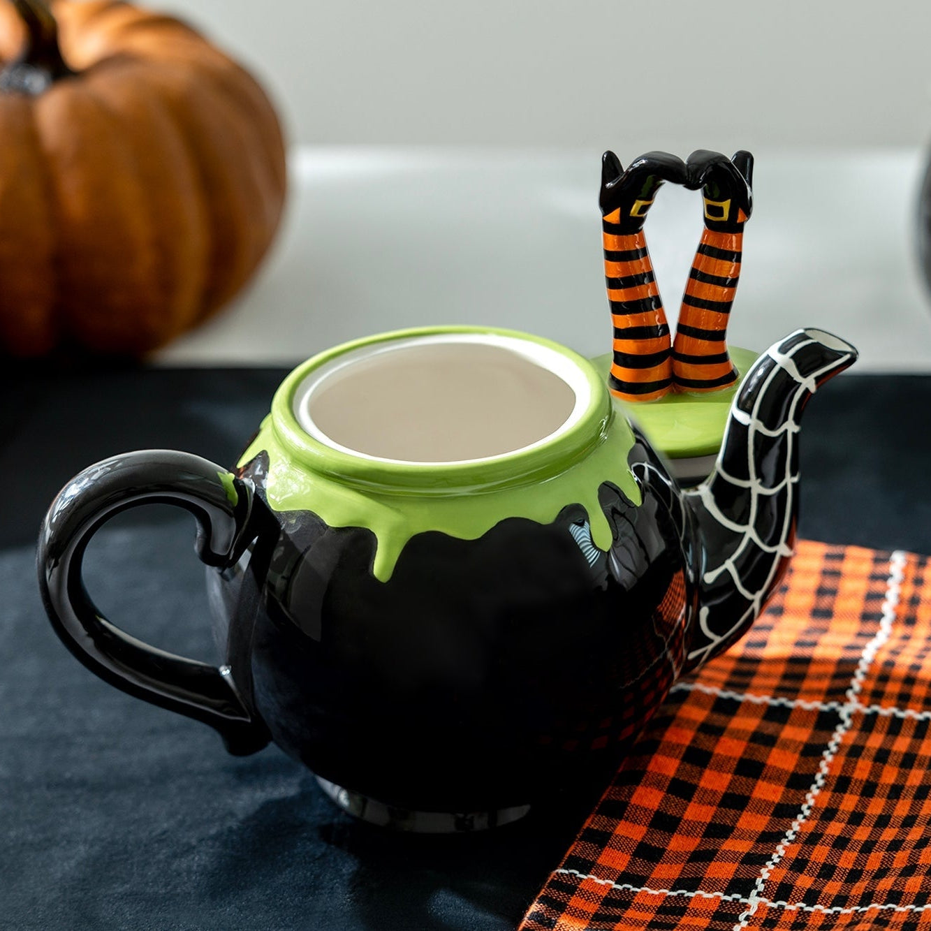 Halloween-themed teapot with green top and black body on a table with pumpkins and checkered cloth.