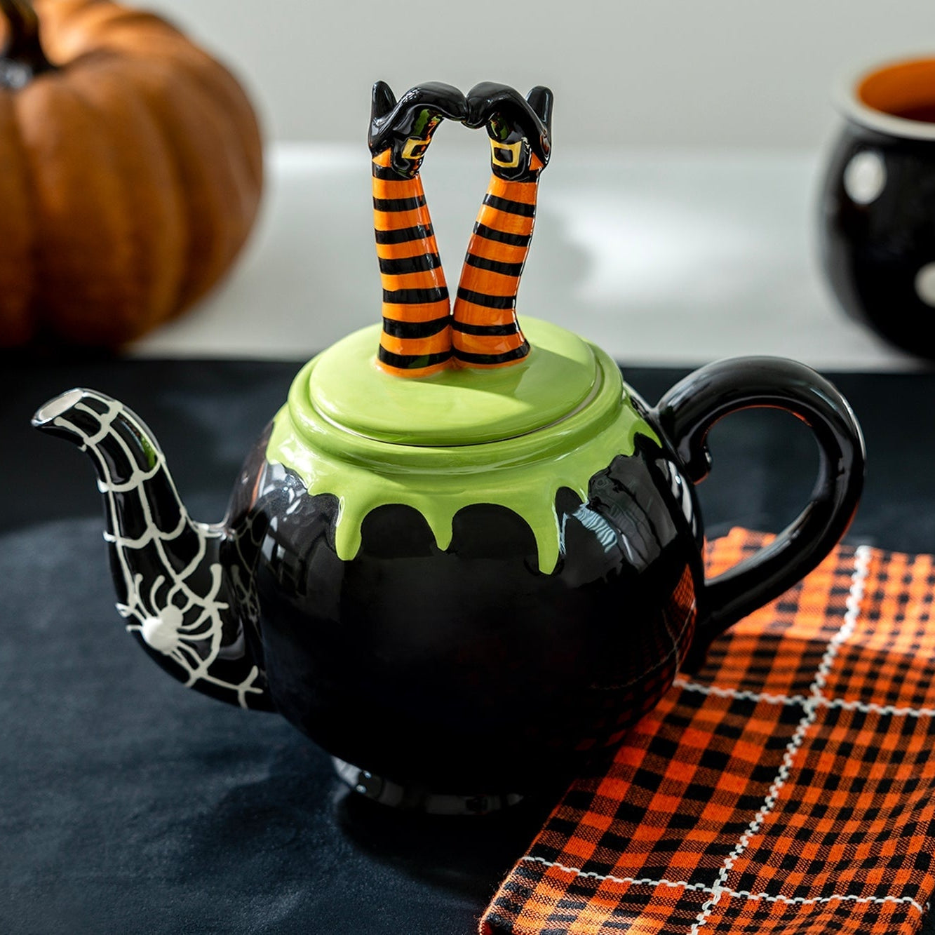 Halloween-themed teapot with witch legs on a table with pumpkins and mugs.