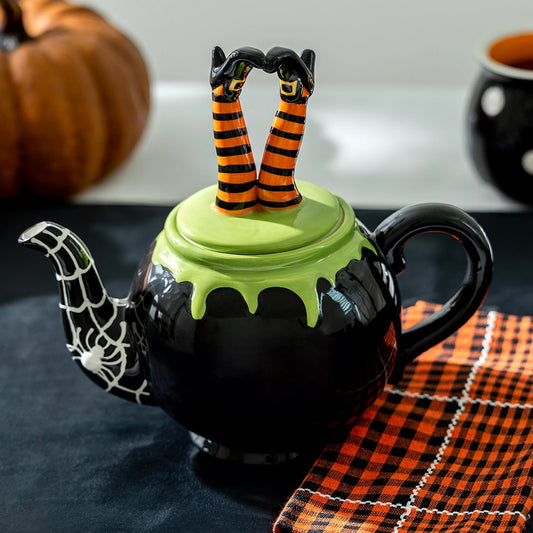 Halloween-themed teapot with witch legs on a table with pumpkins and mugs.